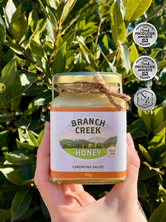 A jar of Local Branch Creek Honey, creamed clover blend honey, held in front of a bush. The label includes the brand name and the phrase 'Cardrona Valley Honey'.